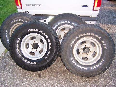 1987 Ford Bronco - Four off-road tires and wheels are displayed in front of a white pickup truck.