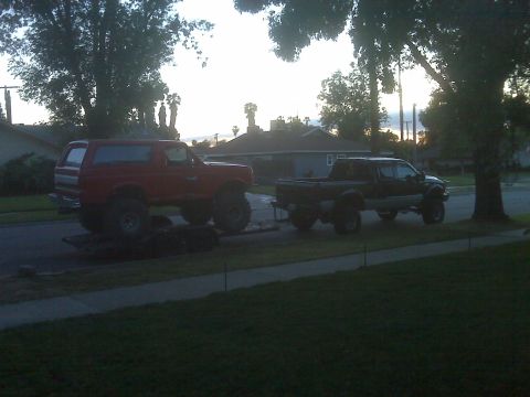 1987 Ford Bronco - Two lifted trucks, a red Bronco and a black F-Series, are parked side-by-side on a street.