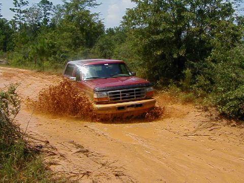 1990 Ford Bronco - This red SUV is kicking up a lot of mud as it drives through a muddy track surrounded by trees.