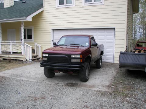 1990 Chevrolet Suburban - A maroon pickup truck with a black bull bar is parked in front of a garage, with another vehicle visible to the right.