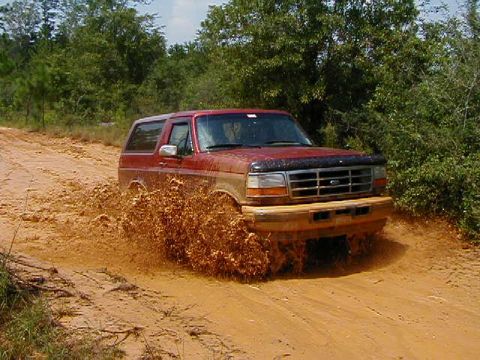 1990 Ford Bronco - A red SUV navigates a muddy trail, kicking up a large spray of dirt.