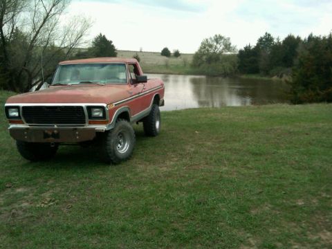 1987 Ford Bronco - A red pickup truck sits on a grassy field next to a calm body of water under a cloudy sky.