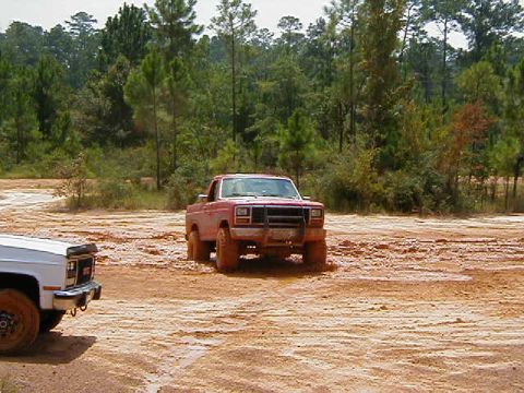 1990 Ford Bronco - Two pickup trucks navigating a muddy off-road course with trees in the background.