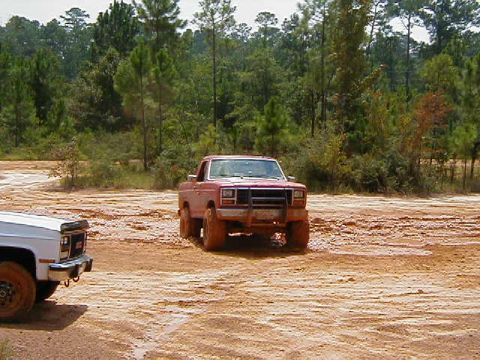 1990 Ford Bronco - Two trucks are shown off-road in a muddy area with trees in the background.