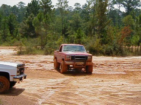 1990 Ford Bronco - Two pickup trucks are seen in a muddy, outdoor area with trees in the background.