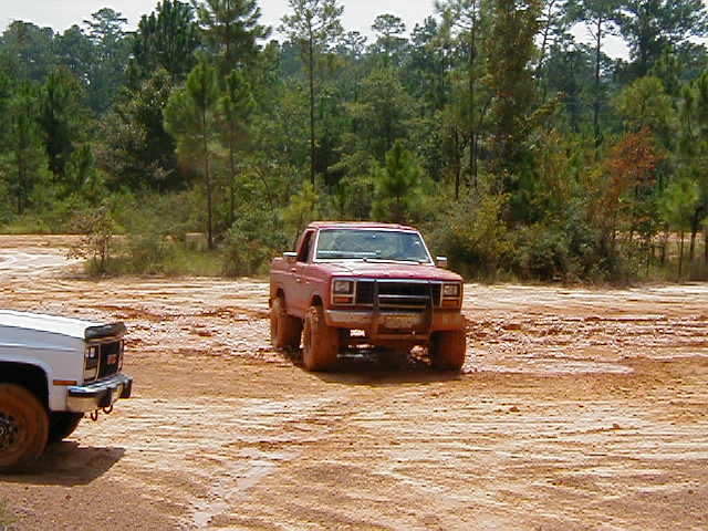 Red pickup truck stuck in mud, front view. White pickup truck to the left