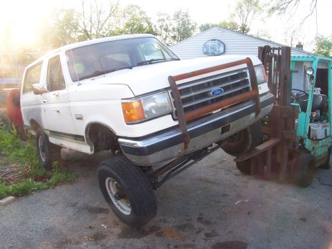 1989 Ford Bronco - White Ford Bronco with a rusty bull bar, lifted front end supported by a forklift.