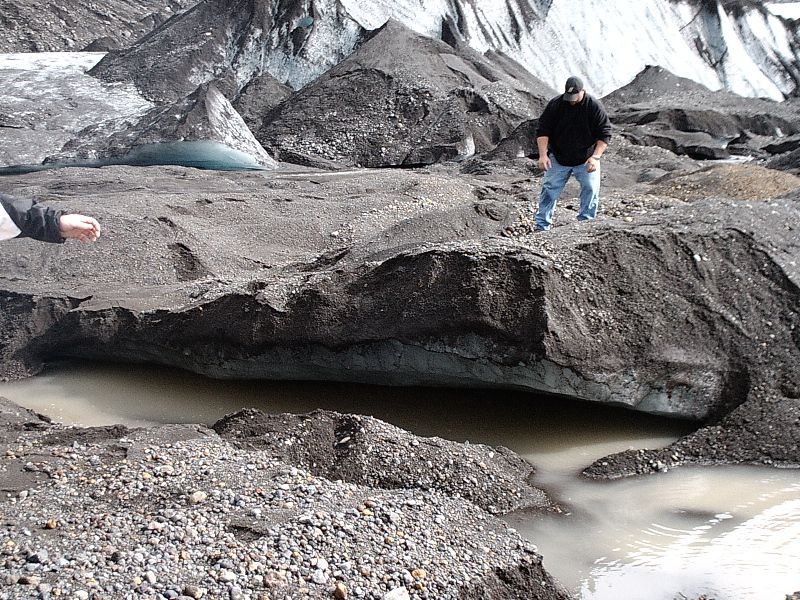 People on a debris-covered glacier near a muddy meltwater pool and ice slopes.
