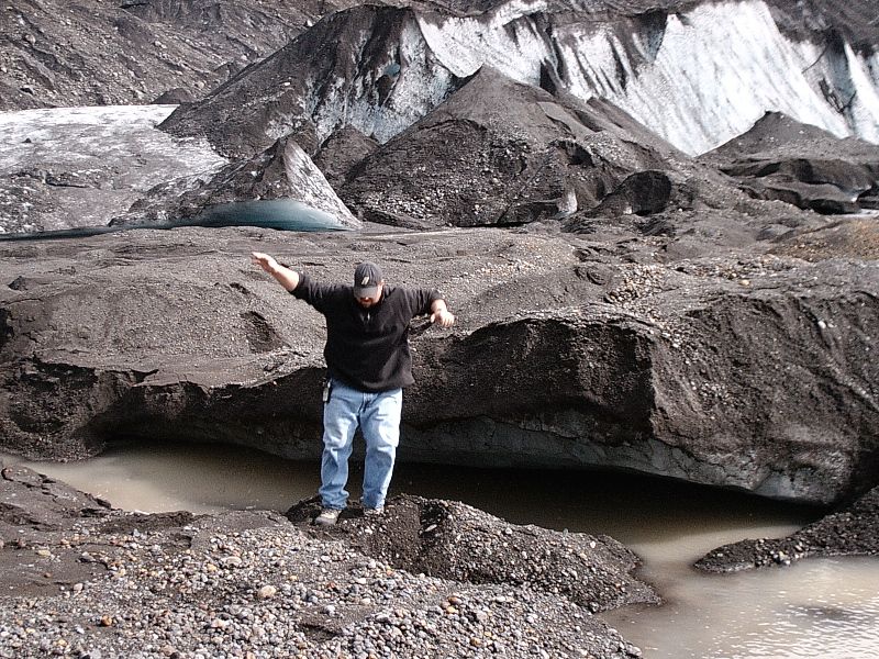 Man standing with arms out on gravel near a debris-covered glacier and murky water.