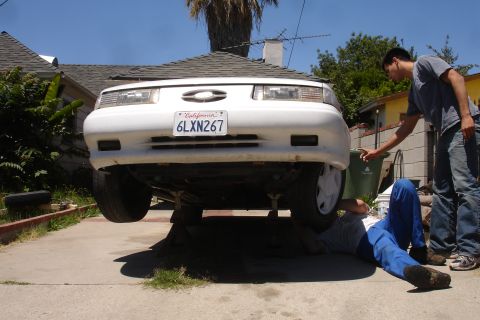 1992 Ford Taurus - Two individuals attend to the underside of a white 1992 Ford Taurus, which is elevated on jack stands.