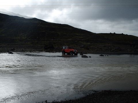 1996 Ford Bronco - An orange Jeep Wrangler navigates a shallow river in a desolate, rocky terrain, with dark hills in the background under an overcast sky.
