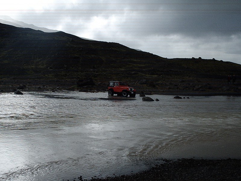 Red SUV fording a wide braided river in a dark, overcast highland landscape
