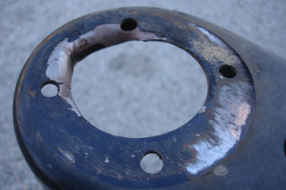 Close-up of a dark, circular automotive part with three bolt holes and chipped paint revealing metal