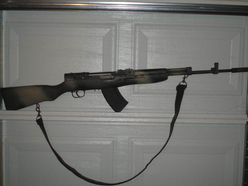 Camouflaged rifle with black magazine and strap hanging on a white garage door