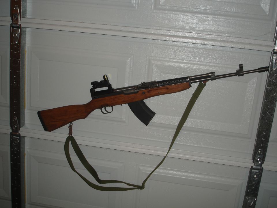 SKS rifle with red dot sight and green sling against a white garage door