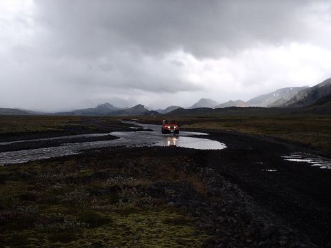 1996 Ford Bronco - A red SUV navigates a watery path through a stark, mountainous terrain under a cloudy sky.