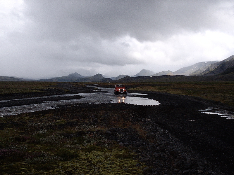 Red SUV fording a river in a dark volcanic landscape under cloudy skies