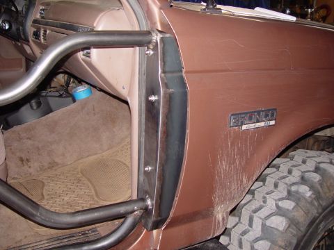 1993 Ford Bronco - Close-up of the driver's door opening on a doorless copper-brown Ford Bronco, showing a bare-steel tube door bar bolted to a fabricated pla…