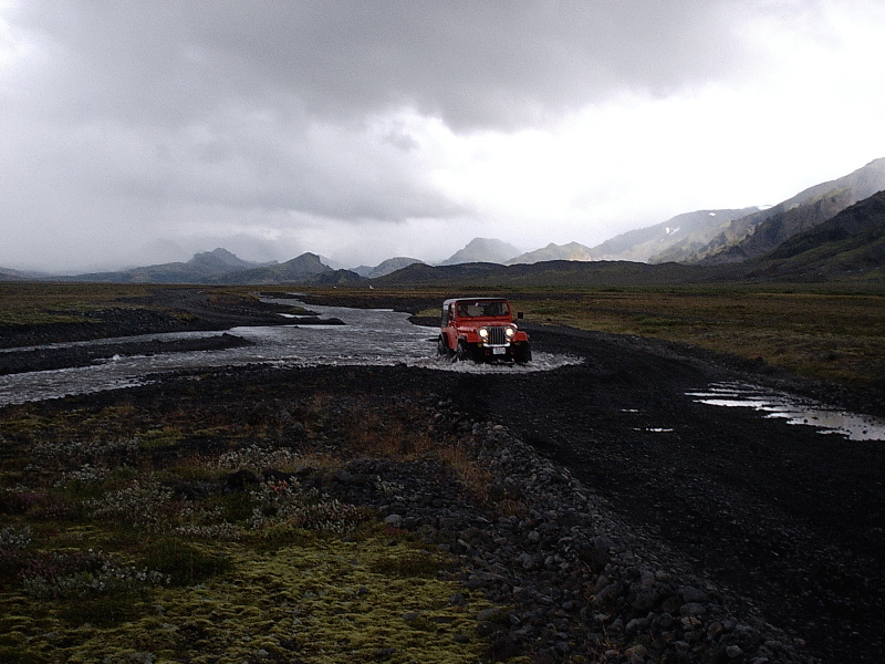 Red SUV with headlights on fording a river across dark gravel amid misty mountains