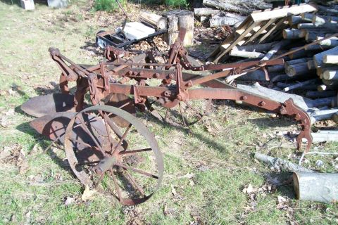 2000 Chevrolet Tahoe - This rusted farm plow, a large spoked wheel, sits in a grassy outdoor setting among logs and debris.
