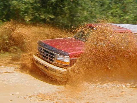 1990 Ford Bronco - A red SUV drives through a muddy puddle, sending up a large spray of dirt.