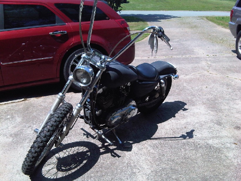 Chopper-style motorcycle with ape hanger handlebars parked on a driveway next to a red SUV