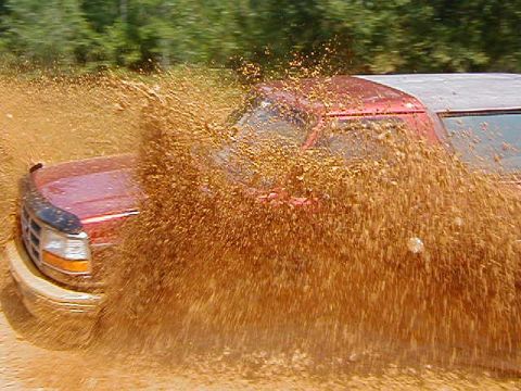 1990 Ford Bronco - A red Ford truck navigates a muddy terrain, sending a large spray of mud into the air.