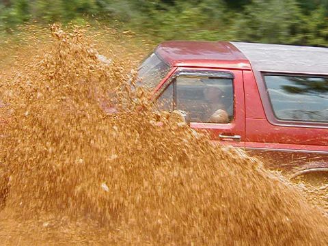 1990 Ford Bronco - A red SUV navigates a muddy terrain, kicking up a large spray of dirt and water.