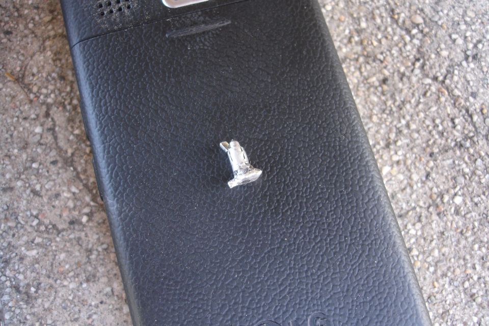 Small, silver, T-shaped metal object with a loop on a black, textured surface, possibly a phone back