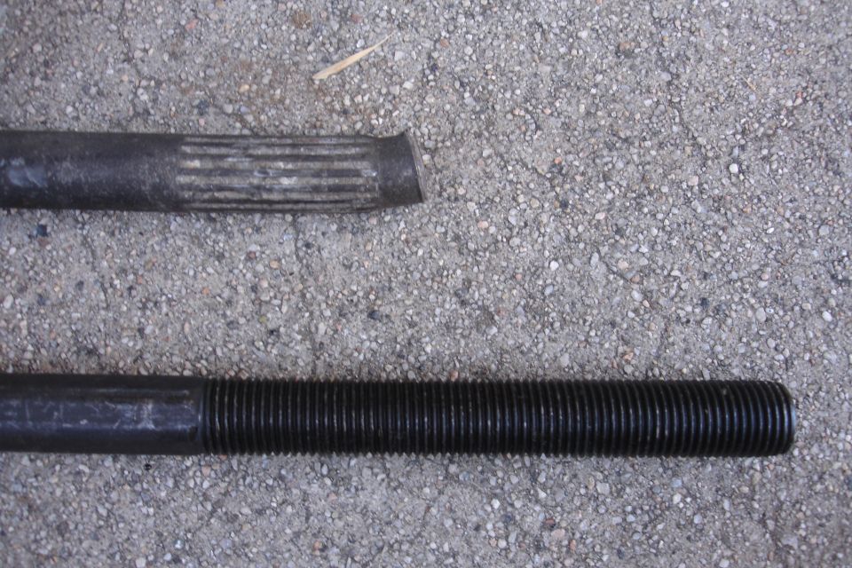 Two metal rods, one with splines and a cut end, the other threaded, lie on a concrete surface