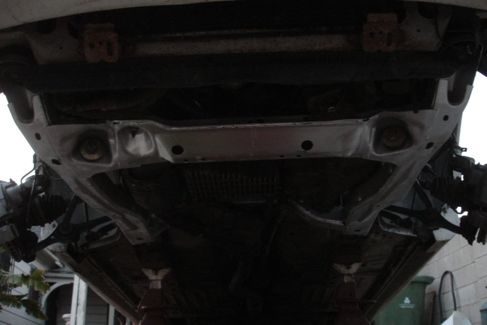 Underside of a car's subframe, supported by jack stands. Some rust is visible on mounting points