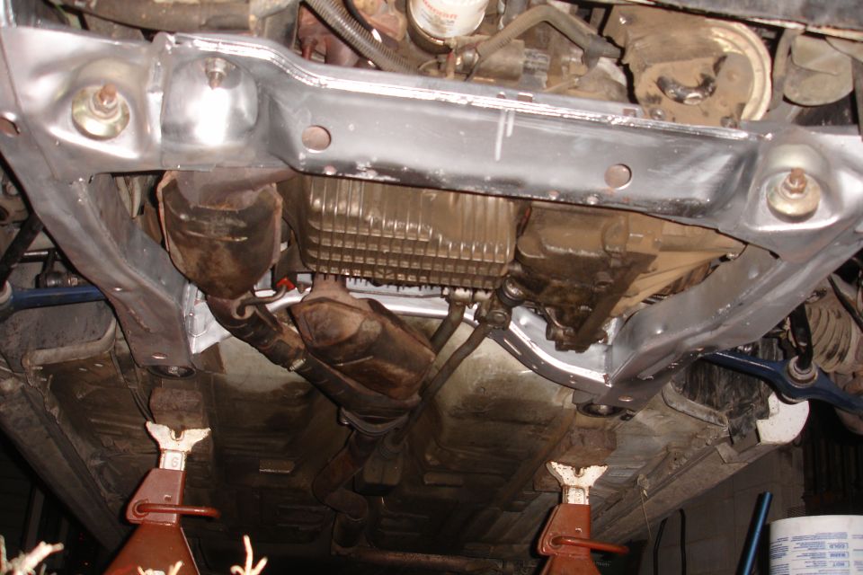Underside view of a car's subframe, exhaust, and transmission components supported by jack stands