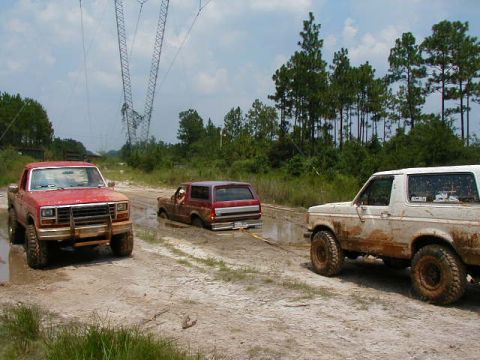 1990 Ford Bronco - Three 4x4 vehicles are stuck in a muddy track with trees and power lines in the background.