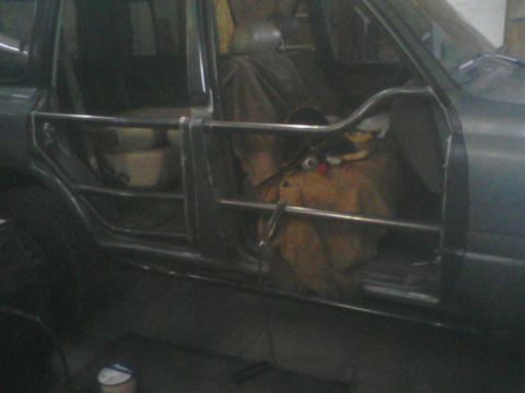 1993 Ford Bronco - Dim garage shot of a bare-metal fabricated tube door mounted on the driver's side of a gray truck, built from bent round tubing with horizo…