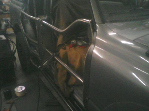 1993 Ford Bronco - Close angled view along the gray fender of a truck toward a shop-built tube door made of bare bent tubing with an angled upper bar and hori…
