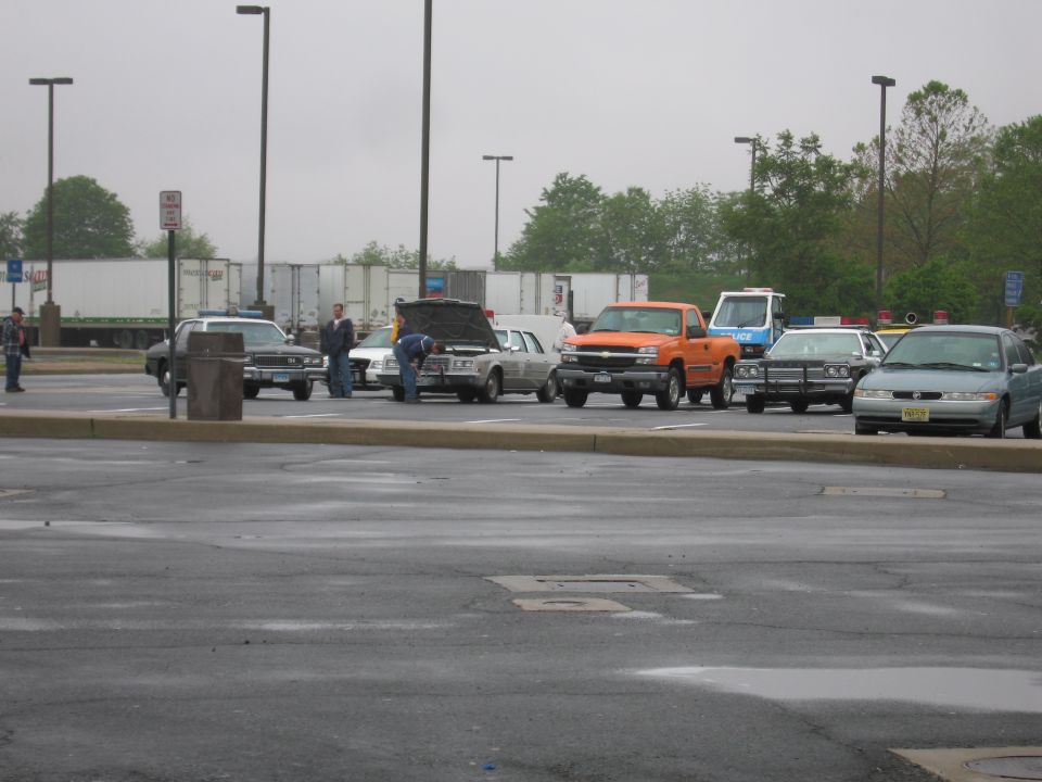 Vintage cars and an orange pickup parked in a wet lot with people nearby