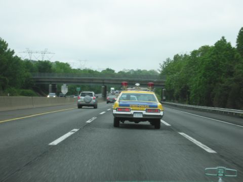 1991 Ford Crown Victoria - A vintage state police car, likely from the 1970s, is seen on the highway with its emergency lights active.