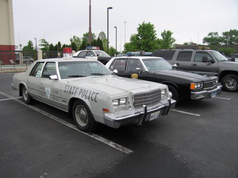 1991 Ford Crown Victoria - Several police vehicles, including a light-colored sedan with 'STATE POLICE' markings, are parked in a lot.