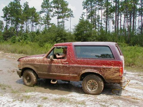 1990 Ford Bronco - A muddy red Ford Bronco drives through a dirt track with trees in the background.