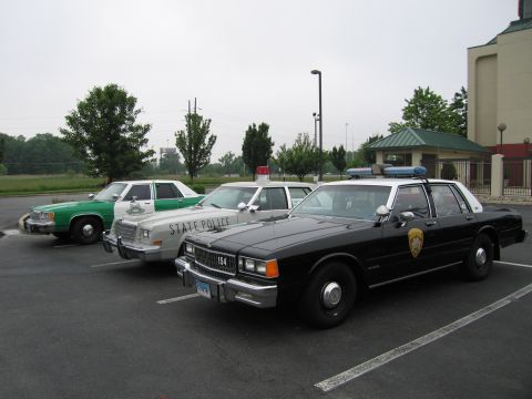 1991 Ford Crown Victoria - Three police cars, including a black one with a badge and number 154 on the grille, are parked in a lot.
