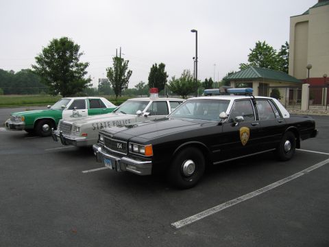 1991 Ford Crown Victoria - Three police cars, including a marked state police vehicle, are parked in a lot under an overcast sky.