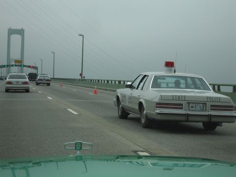 1991 Ford Crown Victoria - A silver police car with "STATE POLICE" on the back is seen from behind on a bridge, with other vehicles and a large suspension bridge in t…