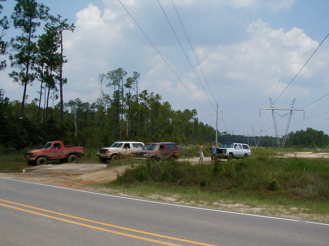 Group of mud-covered trucks and an SUV parked on a dirt shoulder beside a paved road under power lines