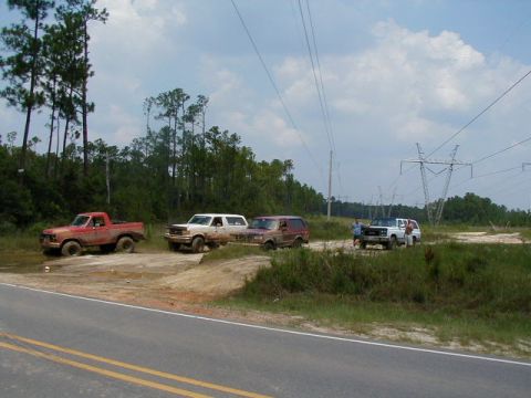 1990 Ford Bronco - Several trucks and SUVs are gathered in a dirt and mud area, with tall power line towers visible in the background.