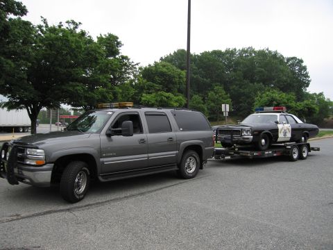 1991 Ford Crown Victoria - A gray SUV tows a trailer carrying a vintage police car with emergency lights on its roof.