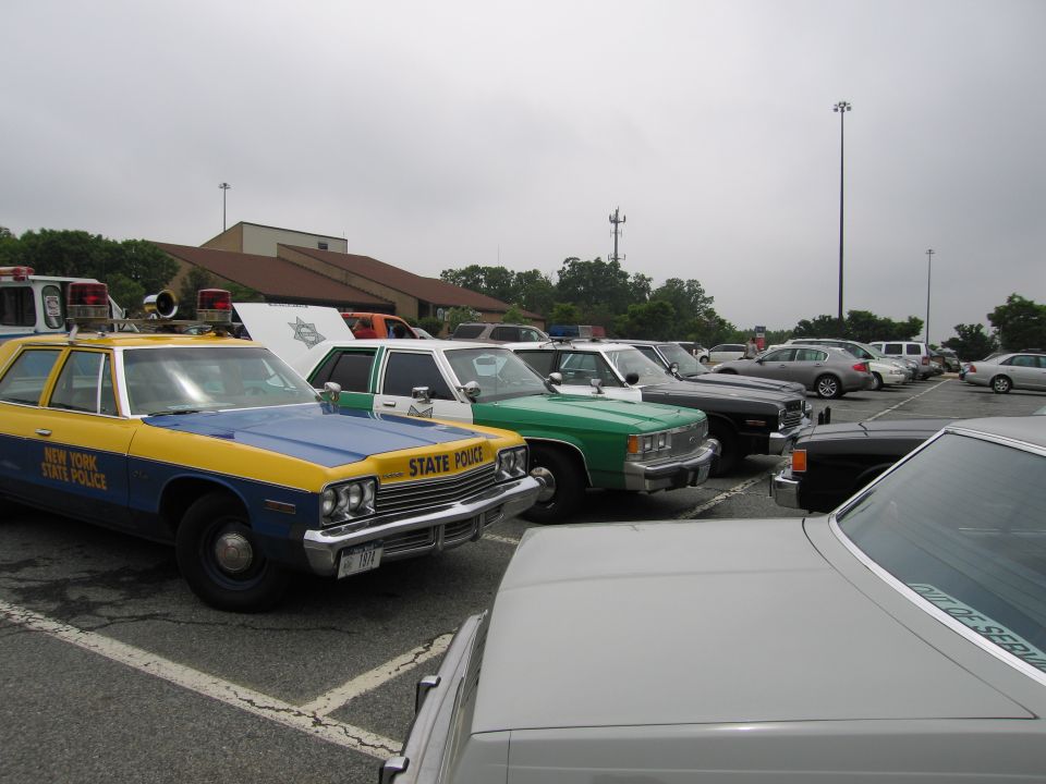 Row of vintage police cars including blue-and-yellow New York State Police sedan and green-and-white car