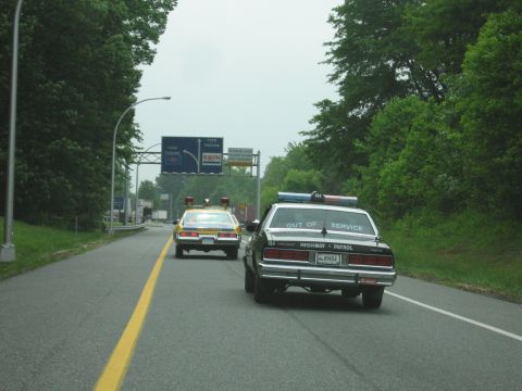 1991 Ford Crown Victoria - Two police cars drive on a highway under an overcast sky.