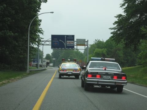 1991 Ford Crown Victoria - Two police cars, one black and one yellow, are seen from behind on a highway under an overpass with signage.