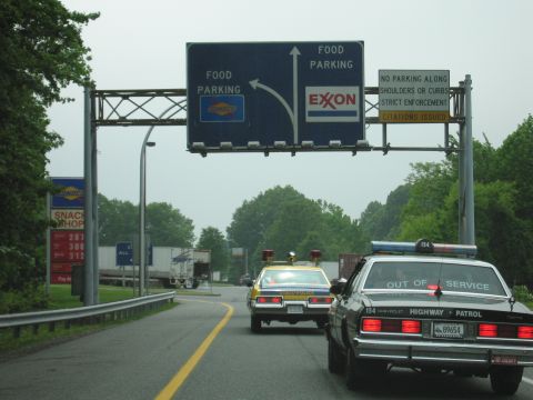 1991 Ford Crown Victoria - Highway patrol cars on the road, with signs for food parking and Exxon ahead.
