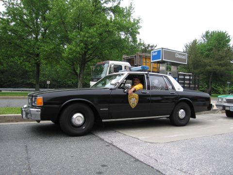 1991 Ford Crown Victoria - A black police car with a uniformed driver is parked at a gas station, with another vehicle visible to the right.